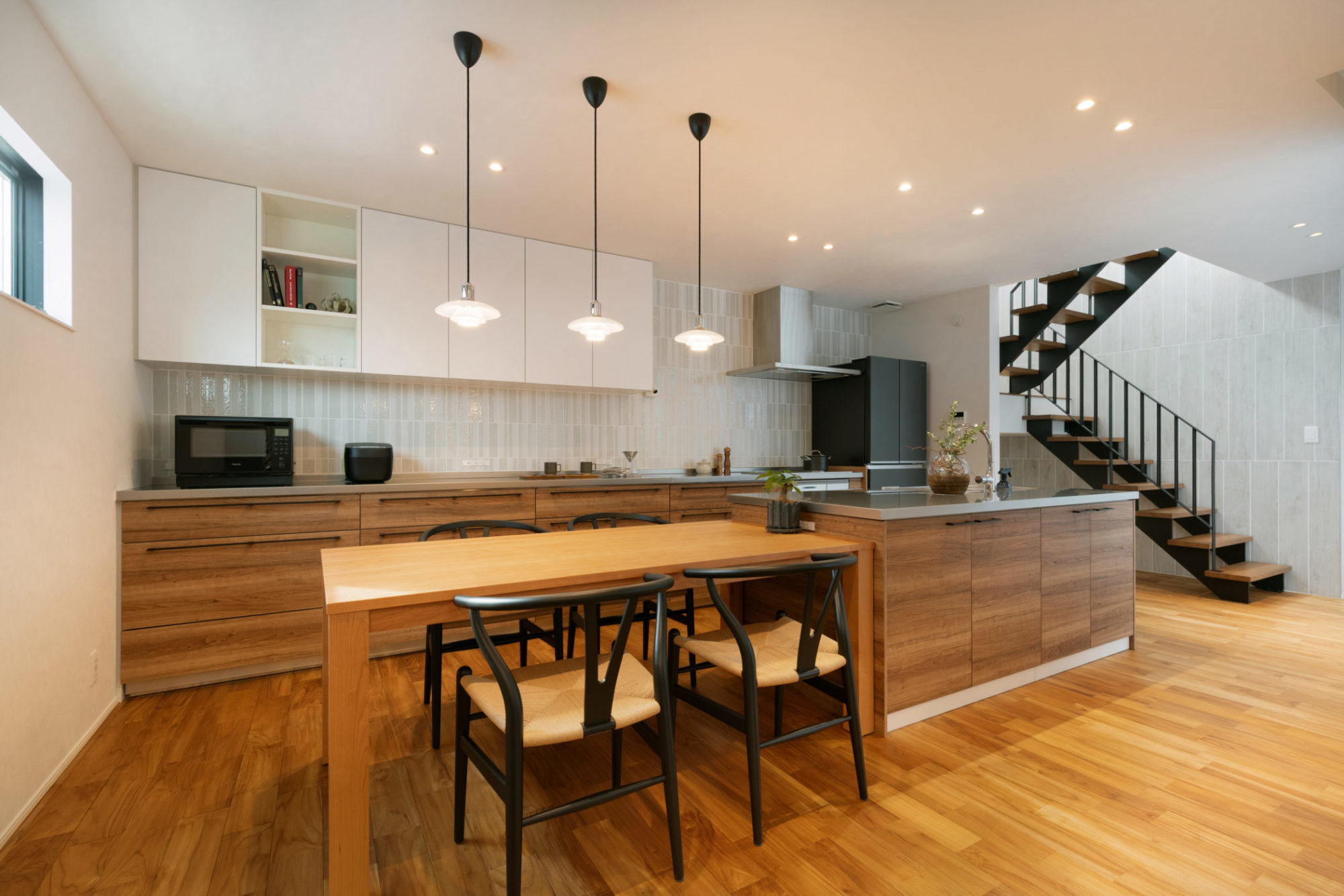 チークの床材とトーンを揃えた木目面材のⅡ型キッチン。ステンレス天板のシャープな質感と、背面壁の質感豊かな白タイルのコントラストが美しい。