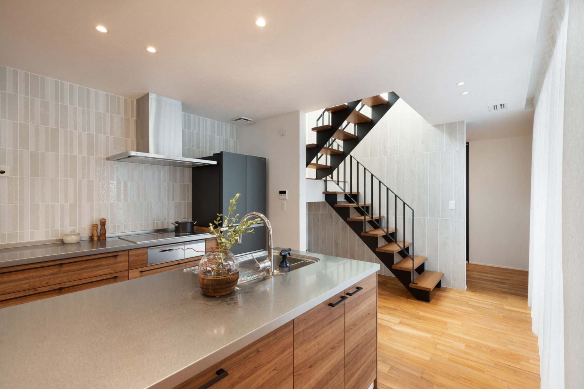 キッチン横に配置された、空間に溶け込むスケルトン階段。木目調タイルの壁面と、繊細なラインが重なり合う意匠性の高い手摺がLDKのアクセントに。